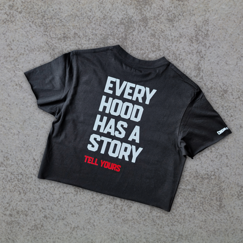 A black cropped t-shirt with bold white text reading 'EVERY HOOD HAS A STORY' and smaller red text reading 'TELL YOURS' underneath, laid flat on a textured gray concrete surface.