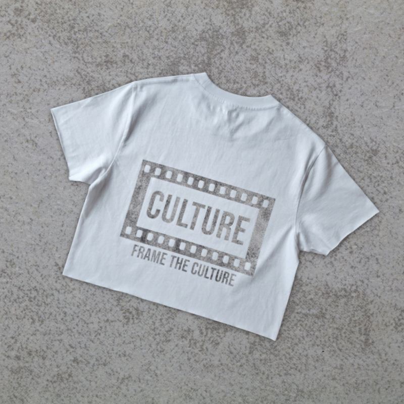 A white short-sleeve crop top t-shirt with a distressed gray graphic on the back featuring a film strip border and the text 'CULTURE FRAME THE CULTURE', laid flat on a textured gray concrete surface.